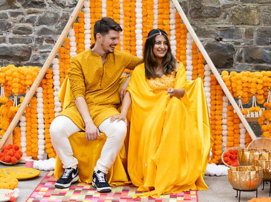 Neary weds sitting together on chairs in The Stables at Plas Dinam Country House. They are dressed in yellow clohing, with orange and white decor behind them. This is their Haldi Ceremony.