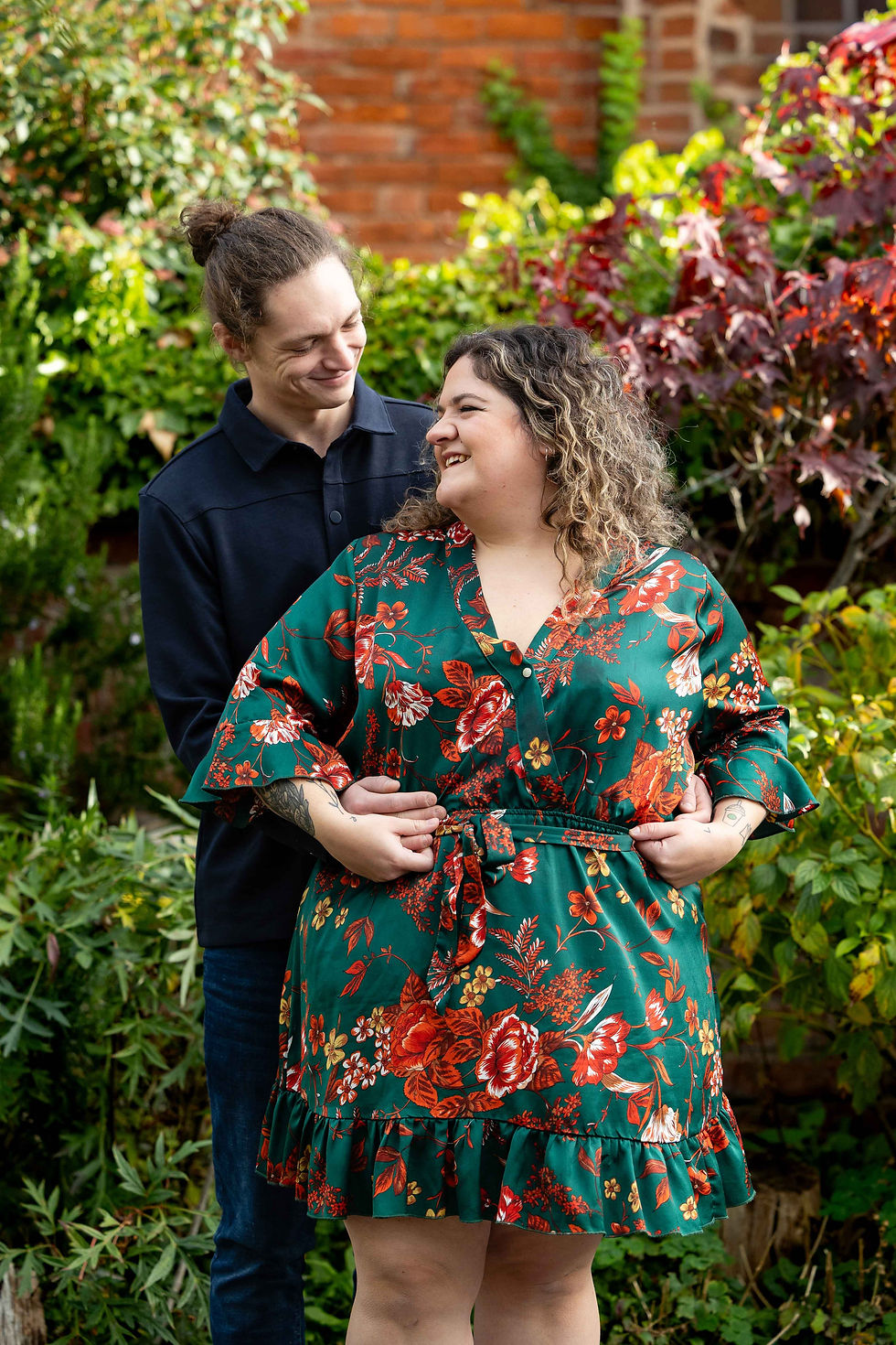 A smiling couple embraces in a garden, the woman in a green floral dress, with red leaves and a brick wall in the background.