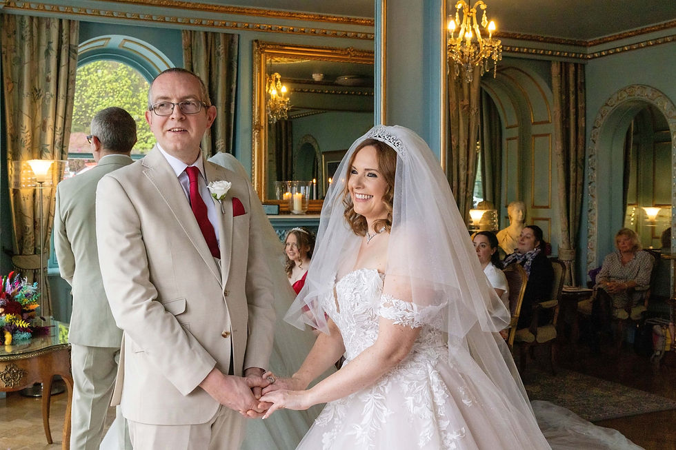 Bride and groom holding hands, smiling in ornate room at portmeirion, with chandeliers and mirrors. Bride in lace gown and veil; groom in beige suit. They are holding hands, taken by Nicki Jones Photography, North Wales wedding photographer.