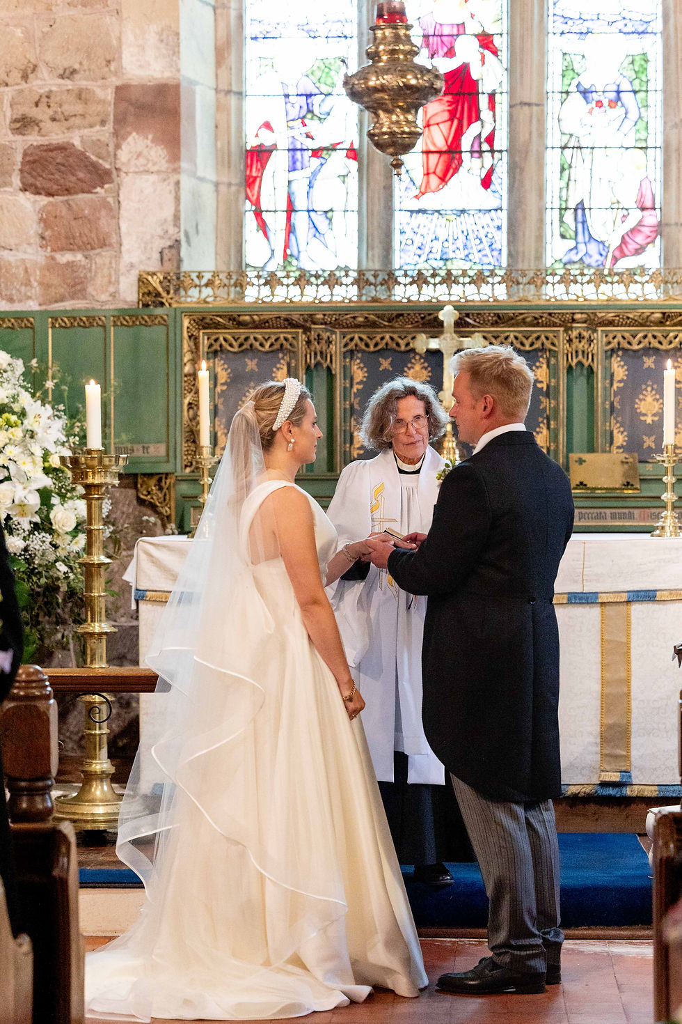 Bride and groom exchange rings in a church wedding ceremony. Priest looks on. Stained glass and candles in the background. Romantic setting.