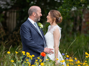 Newly married couple stood facingeach other at Holy Trinity Church in Shrewsbury. They are stood in a patch of wildflowers. The flowers are yellow, red and blue with some wil grass in there too. They are stood facing each other soyou can see a side profile of each of them. They are smiling. The groom holds the brides waist. He wears a blue suit and the bride wears a beautilf lacey dress withshort sleeves