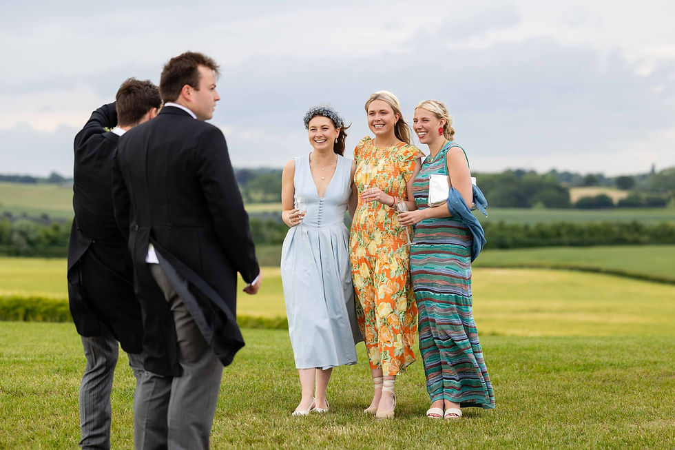 Three women in colorful dresses and two men in suits stand on a grassy field, smiling and holding drinks, with a scenic countryside backdrop.