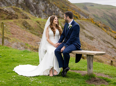 Newly married - couple holding hands while sitting on a bench in the grounds at Nant Gwrtheryn. There are coastal hills in the background. The couple are lookign at each other and smiling. They look happy. The bride wears a beautiful white cloured dress with a long veil,the groom wears a blue suit with a cream button hole.