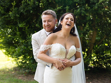 Newly weds standing together and belly laughing. They are surrounded by trees in the gardens at Netley Hall in Shrewsbury.