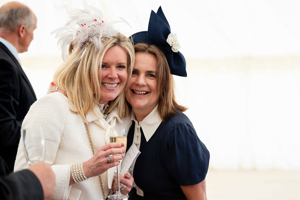 Two women in elegant outfits smile, each wearing decorative hats. One holds a champagne glass. Bright, festive indoor setting.