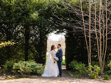 Newly married couple standing and facing each other, holdig hand in the gardens at Netley Hall in Shrewsbury. They are looking at each other and smiling. They are surrounded by greenery and bushes and are stood in an natural arcway in the hedge which frames them. The groom wears a blue suit and cream button hole. The bride wears a beautiful long wedding dress with veil and is holding a bouquet in her right hand.