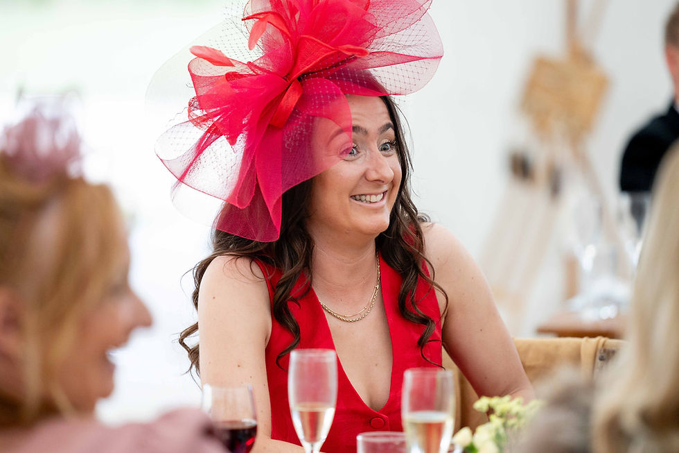 Woman in red dress and large red fascinator smiles at event. Champagne glasses on table. Bright, cheerful atmosphere.