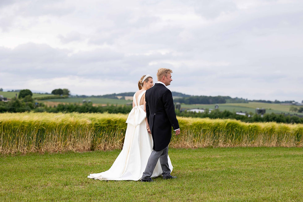 A newly married couple walk hand in hand through a grassy field. She's in a white dress, he's in a black suit. Green fields stretch behind them.