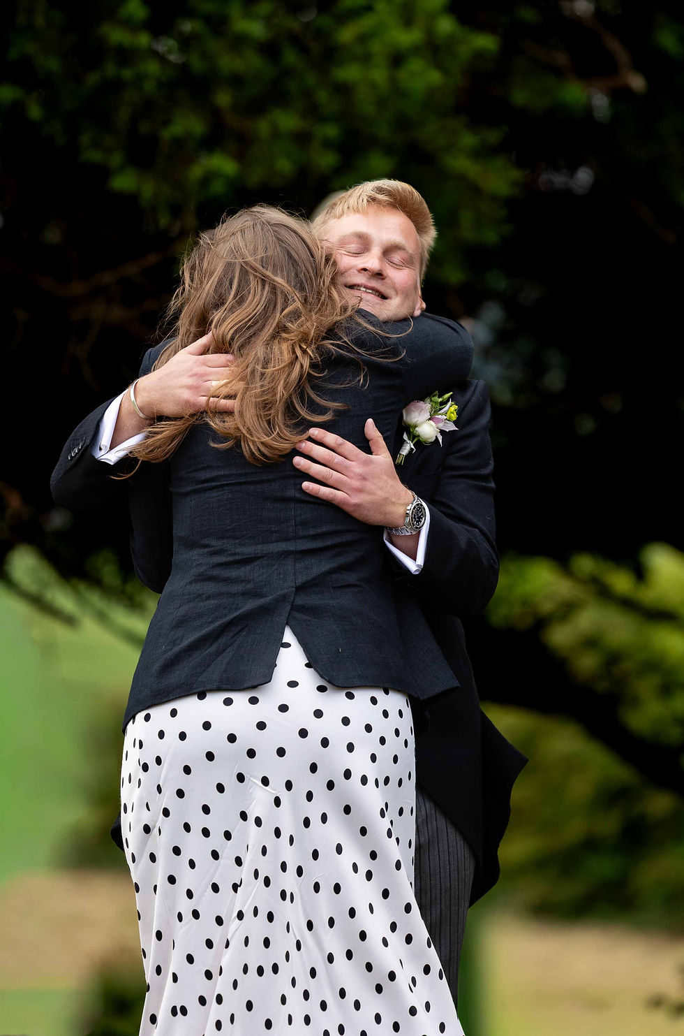 Two people embracing warmly outdoors. One wears a polka-dot skirt, the other a suit with a boutonniere. Green foliage in the background.