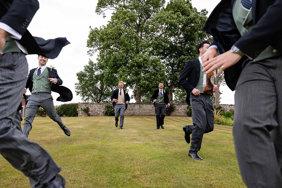 Groomsmen in formal attire run across a grassy lawn, trees and a stone wall in the background. Lively mood.