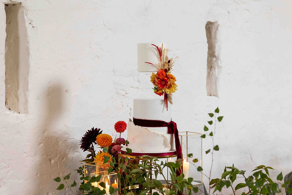 Elegant white cake with red and orange flowers and ribbon, set against a textured white wall with green leaves and candles below.