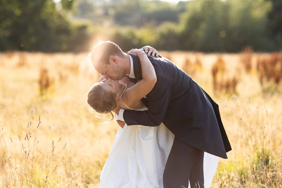 A couple kissing in a sunlit field, the man in a suit dips the woman in a white gown. It is a warm, romantic setting with golden grass. Taken by Shropshire wedding photographer, Nicki Jones Photography.