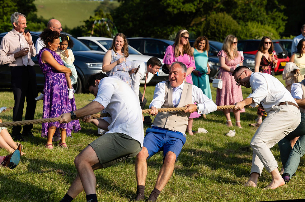 Wedding guests in colourful attire playing tug-of-war on grass, laughing and enjoying a sunny day. Onlookers in the background, standing in a field.