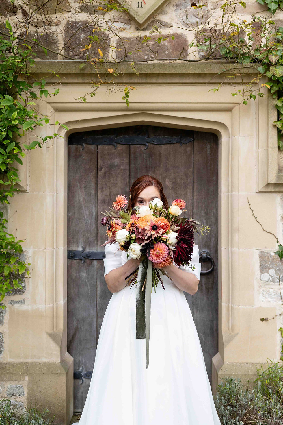 Bride in white dress holds colorful bouquet, standing in front of rustic wooden door framed by ivy and stone, creating a romantic scene.