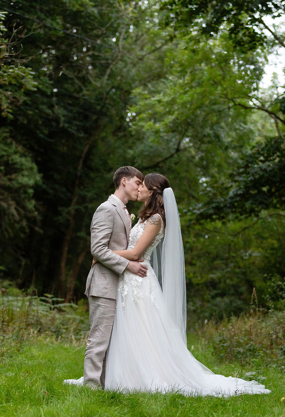 Bride and groom kiss in a lush green forest, she in a white gown with veil, he in a beige suit. Romantic and serene setting.