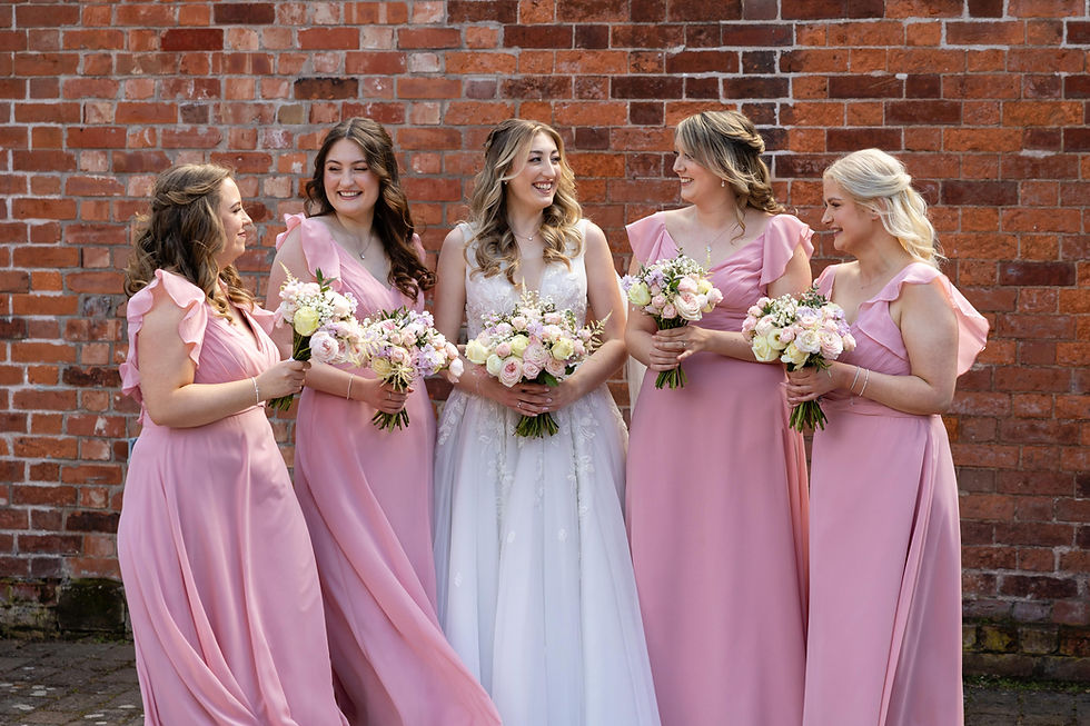 Bride and four bridesmaids in pink dresses holding bouquets, they are smiling in front of a rustic brick wall at neatly hall in Shrewsbury. It is a bright and joyful atmosphere.