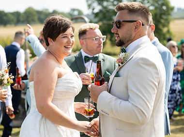 Newly married couple, holding hands while smiling. They are bothholding drinks and are surrounded by theiur gusts. They look happy. The bride is wearing a white strapless dress and the groom a cream suit.