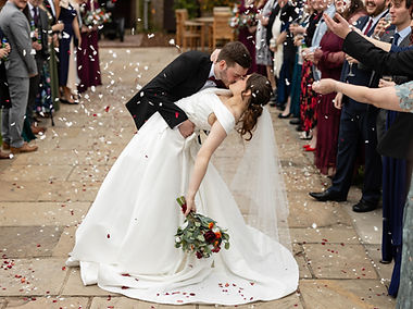 Bride and groom doing a dip and kiss in teh gardens at Rowton Castle. They are surroudned by their guests who are throwing confetti. The confetti is in deep reds and creams. The bride wears a white dress with a long veil. The groom si wearing a black tuxedo and bow tie.