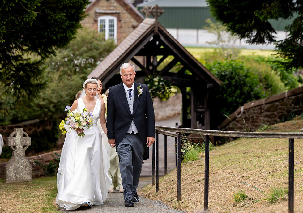 Bride in white gown holds bouquet, walking with older man in formal attire. Stone building and greenery in the background.