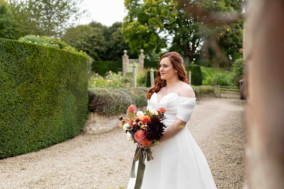 Bride in a white dress holding a colorful bouquet stands in a garden with green hedges and trees, exuding a serene and joyful mood.