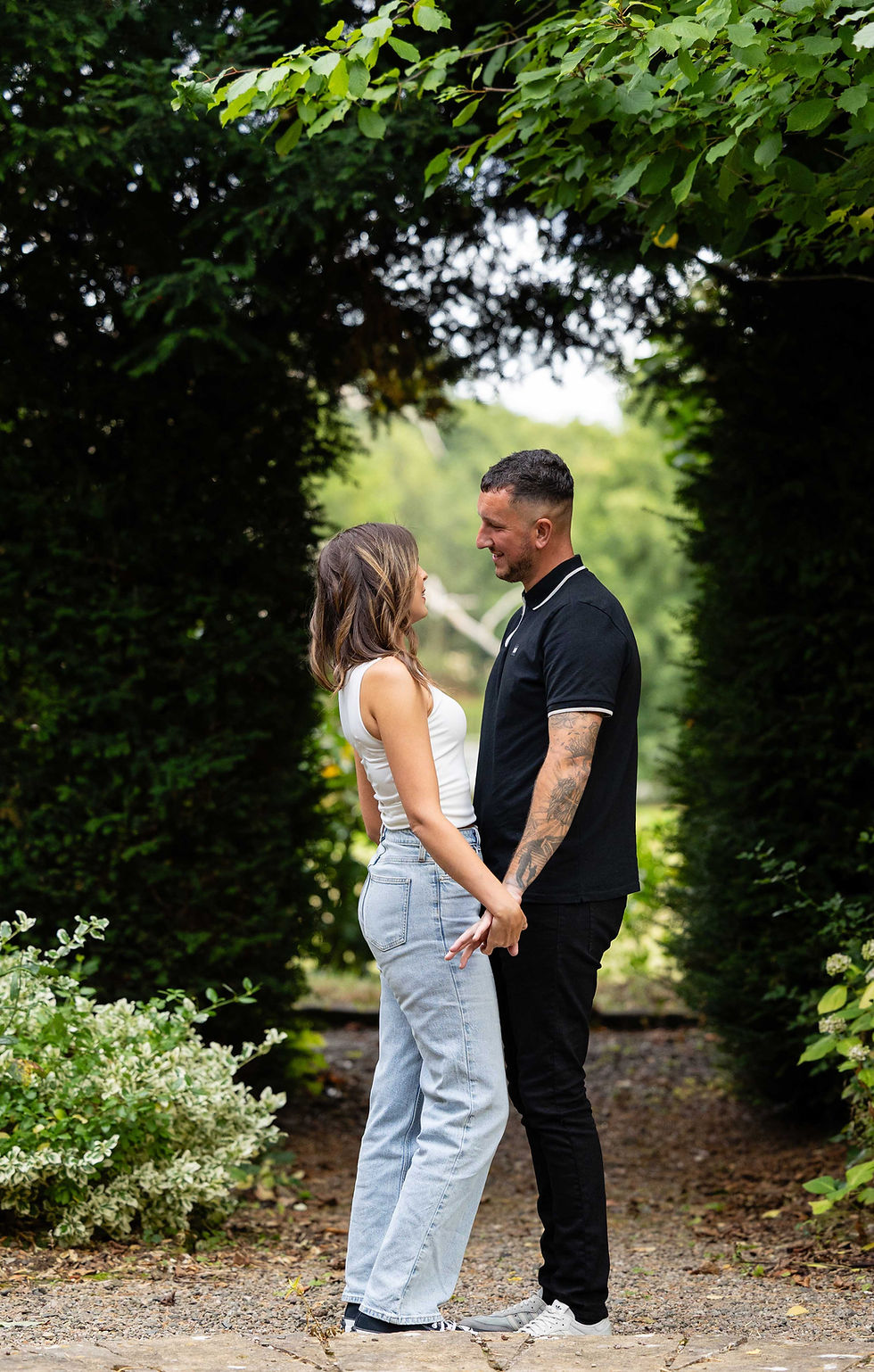 A couple holds hands and gazes at each other, standing on a garden path surrounded by lush greenery in the grounds at Netley Hall in Shrewsbury. They're smiling and relaxed. Taken by Nicki Jones Photography, Shrewsbury wedding photographer.