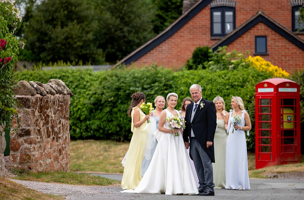 Bride and bridesmaids in pastel dresses pose with an older man in a garden setting. Red phone booth in background, cheerful mood.