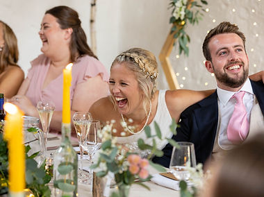 Bride belly laughing while sitting at a table during her wedding speeches at The Courtyard Venue in oswestry. She sits next to her new husband who is wearing a blue suit and a pink tie and her bridesmaid is in th background wearing a pink dess. Sparkly fairy lights are in the bakground.