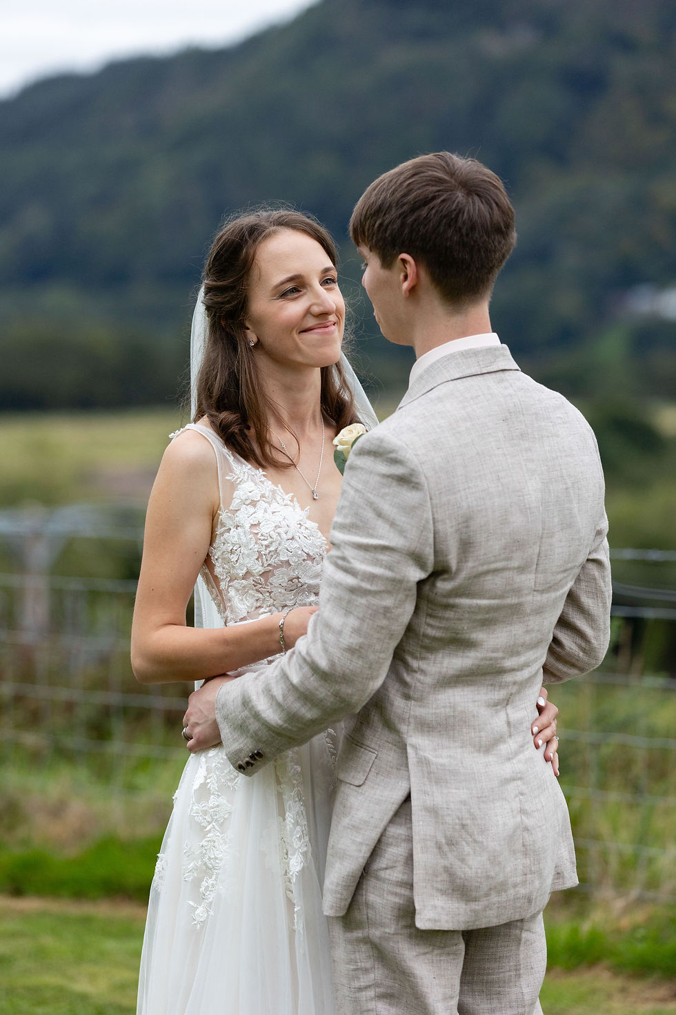 A couple in wedding attire embraces outdoors, smiling at each other with a mountainous landscape in the background.