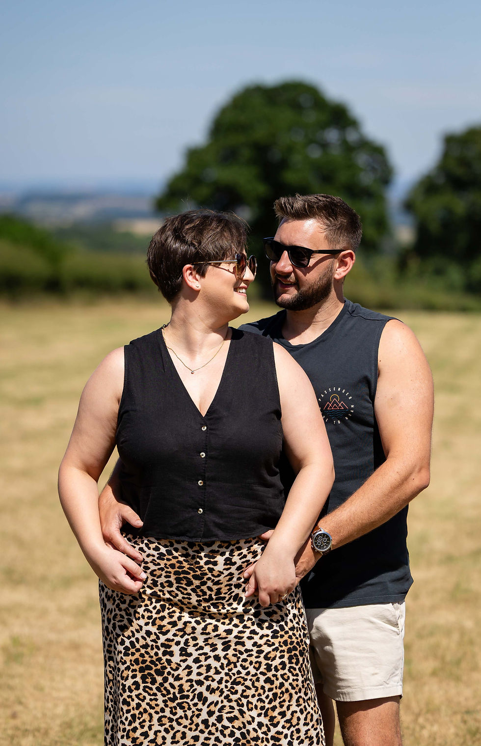 A couple in sunglasses smiles at each other in a sunny field during their pre wedding photoshoot. She wears a black top and leopard skirt, and he has a black tank and shorts. They ;look happy and are smiling at each other. Taken by Nicki Jones Photography, Shropshire wedding photographer.