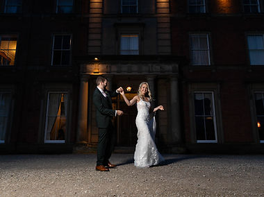 Brieand groom holding hands outside of Netley Hall in Shrewsbury. It is dark, the couple are lit by a singularlight and theyare both smiling.