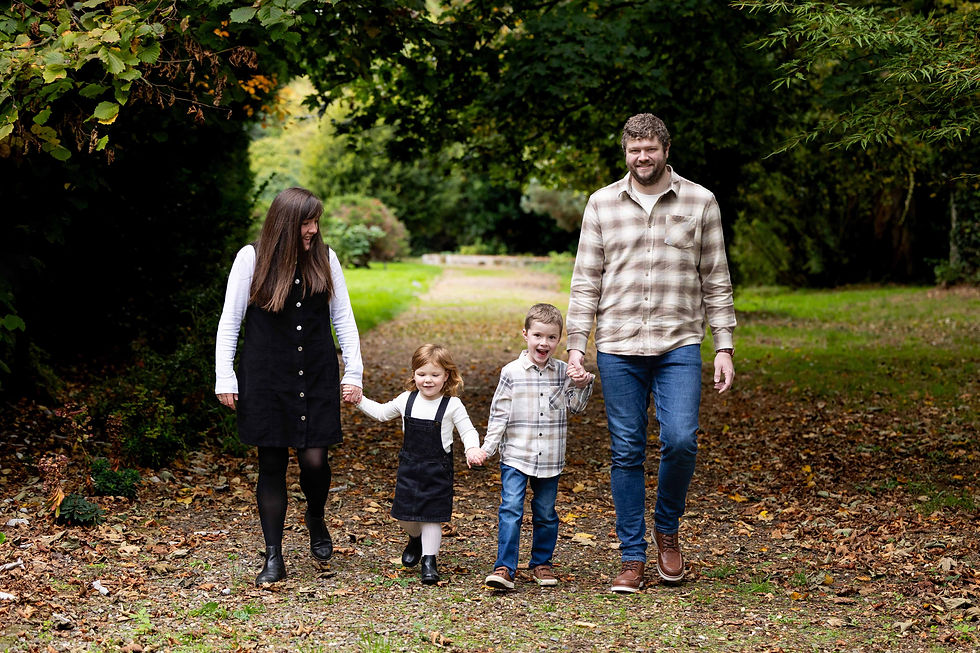 Family of four walking hand-in-hand on a leaf-strewn path at Netley Hall in Shrewsbury. Parents are smiling at their two children. There are lush green trees surrounding them in a serene setting.