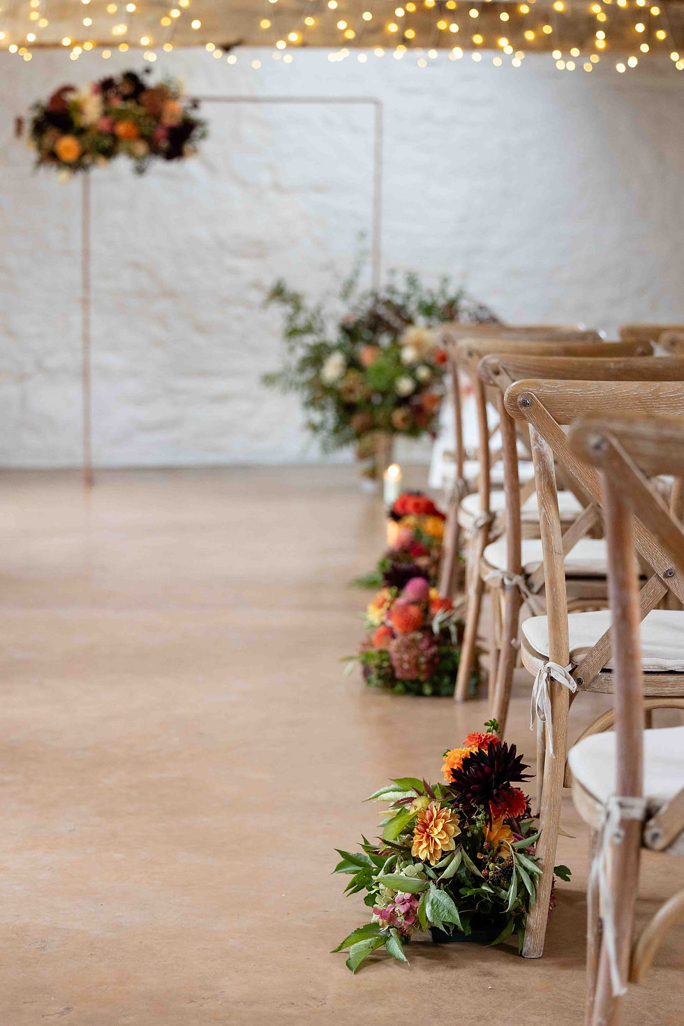 Elegant rustic wedding aisle with wooden chairs, vibrant floral arrangements, fairy lights, and a white brick wall, creating an intimate mood for a wedding ceremony at the Courtyard Venue in Oswestry, Shropshire.