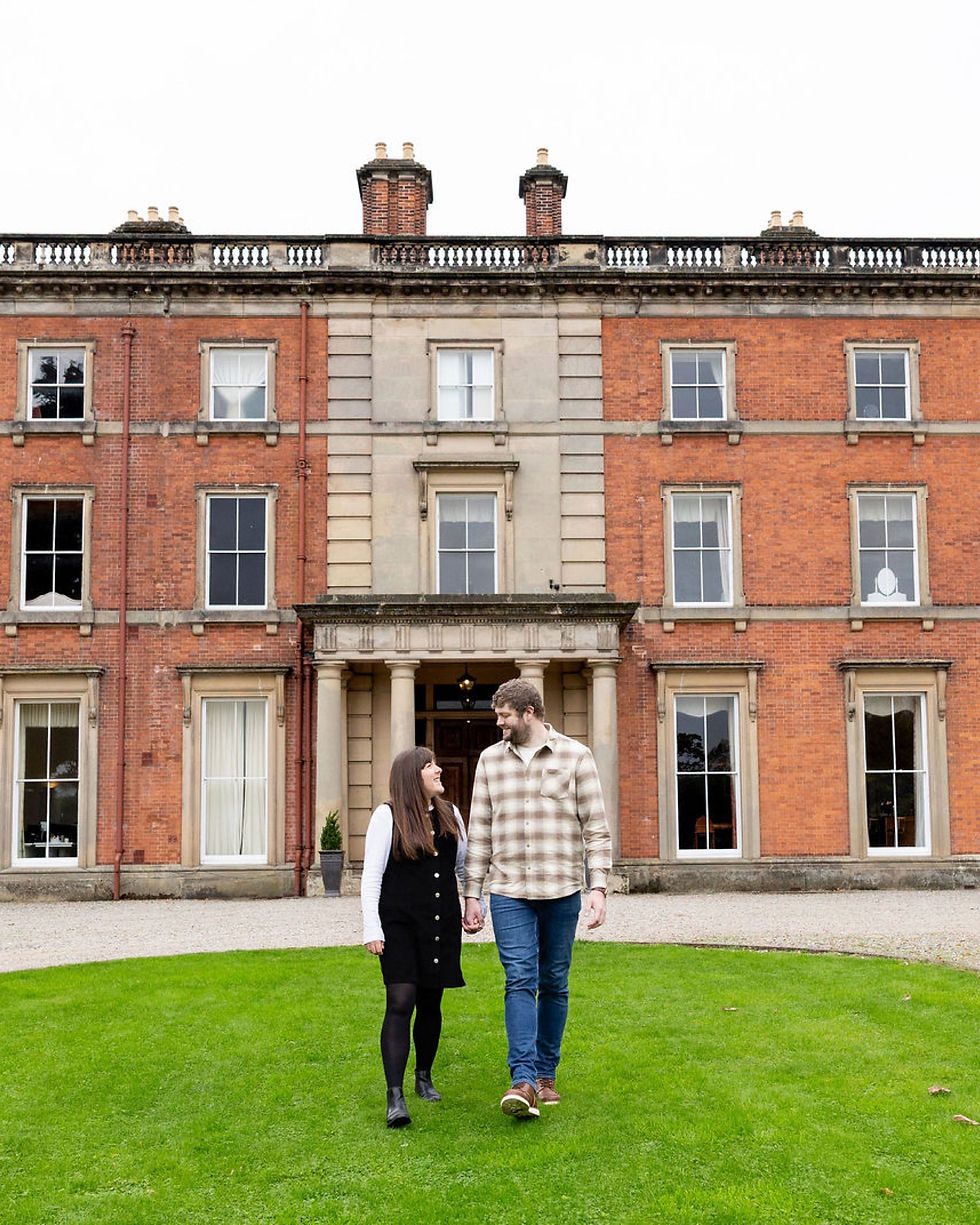 A couple walk hand in hand on green grass in front of a grand brick building at Netley hall in Shrewsbury. It has tall windows, creating a warm, peaceful atmosphere.