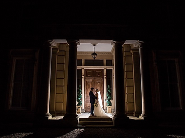 Newly married couple standing and fancing each other in betweentwo coloumns outside the front of Netley Hall in Shrewsbury. It is nighttime, and it isdark. The couple are silhoutted against the front doors.