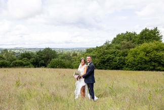 Newly married couple standing a in field, embracing. They are surrounded by a field of grass with trees in the background. 