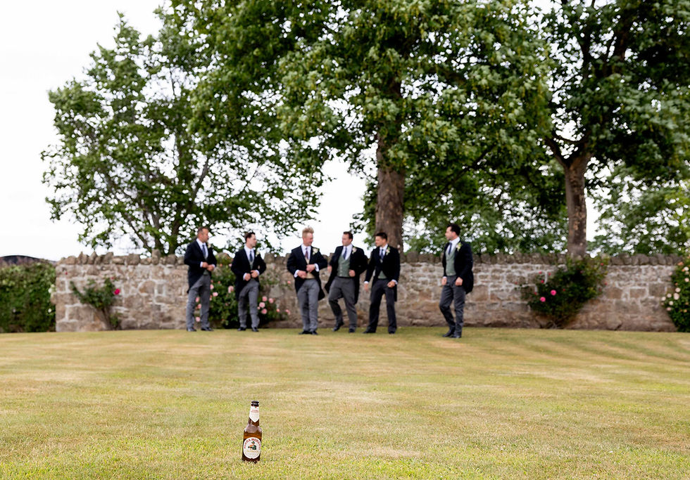 A group of men in suits walk in a garden, blurred in the background; a beer bottle is in focus on the grass in the foreground.