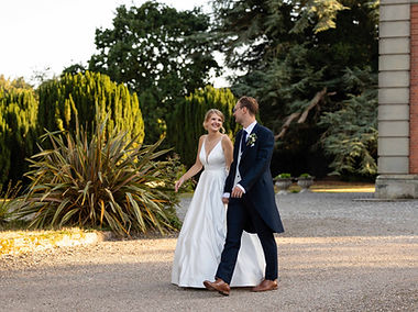 A newly married couple,walking hand in hand hrough the gardens at Netley Hall in Shrewbsury. The bride is wearing a long white dress, the groom a bliue suit. They look happy, and it is a candid photo.