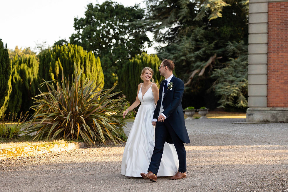 Bride in a white dress and groom in a navy suit walk through the gardens at Netley Hall in Shrewsbury, smiling at each other. The background features lush greenery and red brick wall.