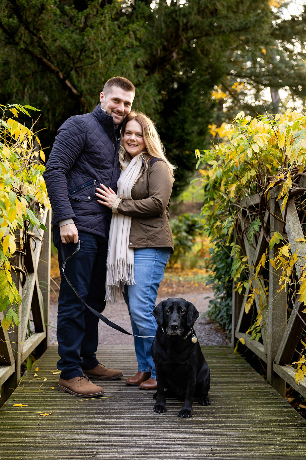 Couple hugging on a leafy bridge with a sitting black labrador. They're dressed warmly, exuding happiness amidst lush greenery and autumn hues.