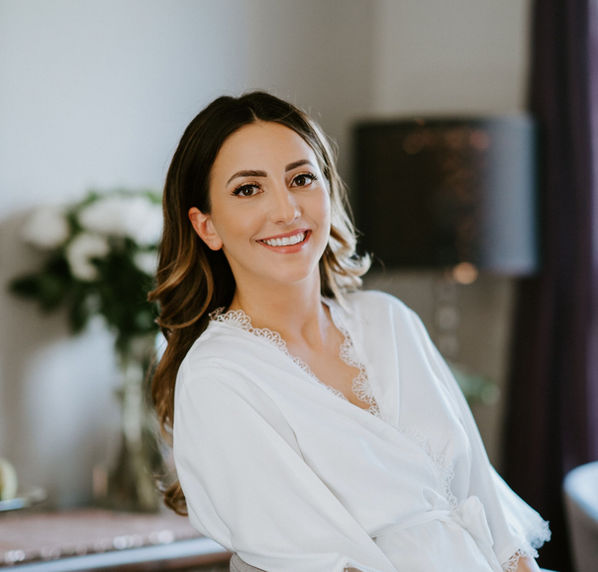 Smiling woman in her white robe while getting her bridal makeup done for her wedding. She has a beautiful blowout and style from Brooklin Mane Hair Studio