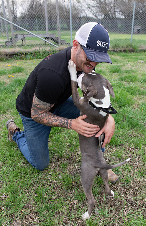 A kneeling man in a baseball hat hugging a small grey and white puppy