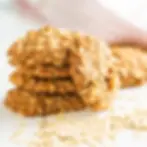 A neat stack of golden raisin-free oatmeal cookies on a white wooden surface with loose oats nearby