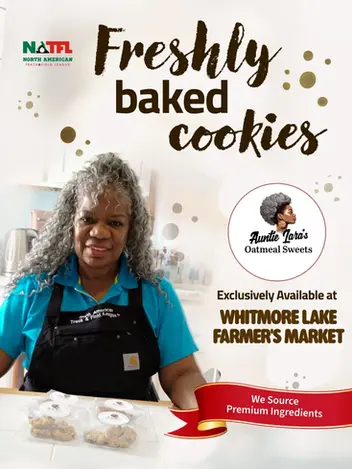 A smiling woman wearing an apron stands behind packages of Auntie Lara's Oatmeal Sweets, exclusively available at the Whitmore Lake Farmer's Market.