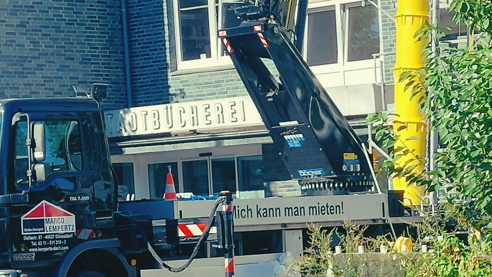 Baukran-LKW vor einer Bücherei; gelbes Rohr rechts; Aufschrift Mich kann man mieten!