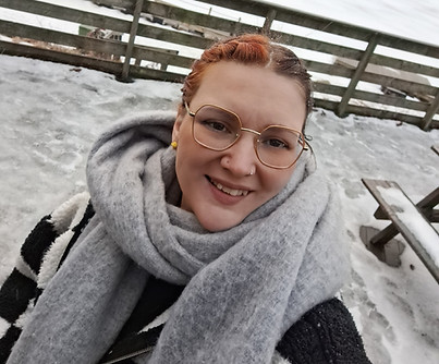 Photo of Emma. Her hair is in braids. She wears gold rimmed glasses. She has a thick scarf around her neck which is grey. Behind her the scenery is of snow.
