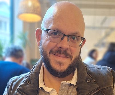This is a photo of Jason. He is smiling for the camera. He is bald with a short beard and wears glasses.