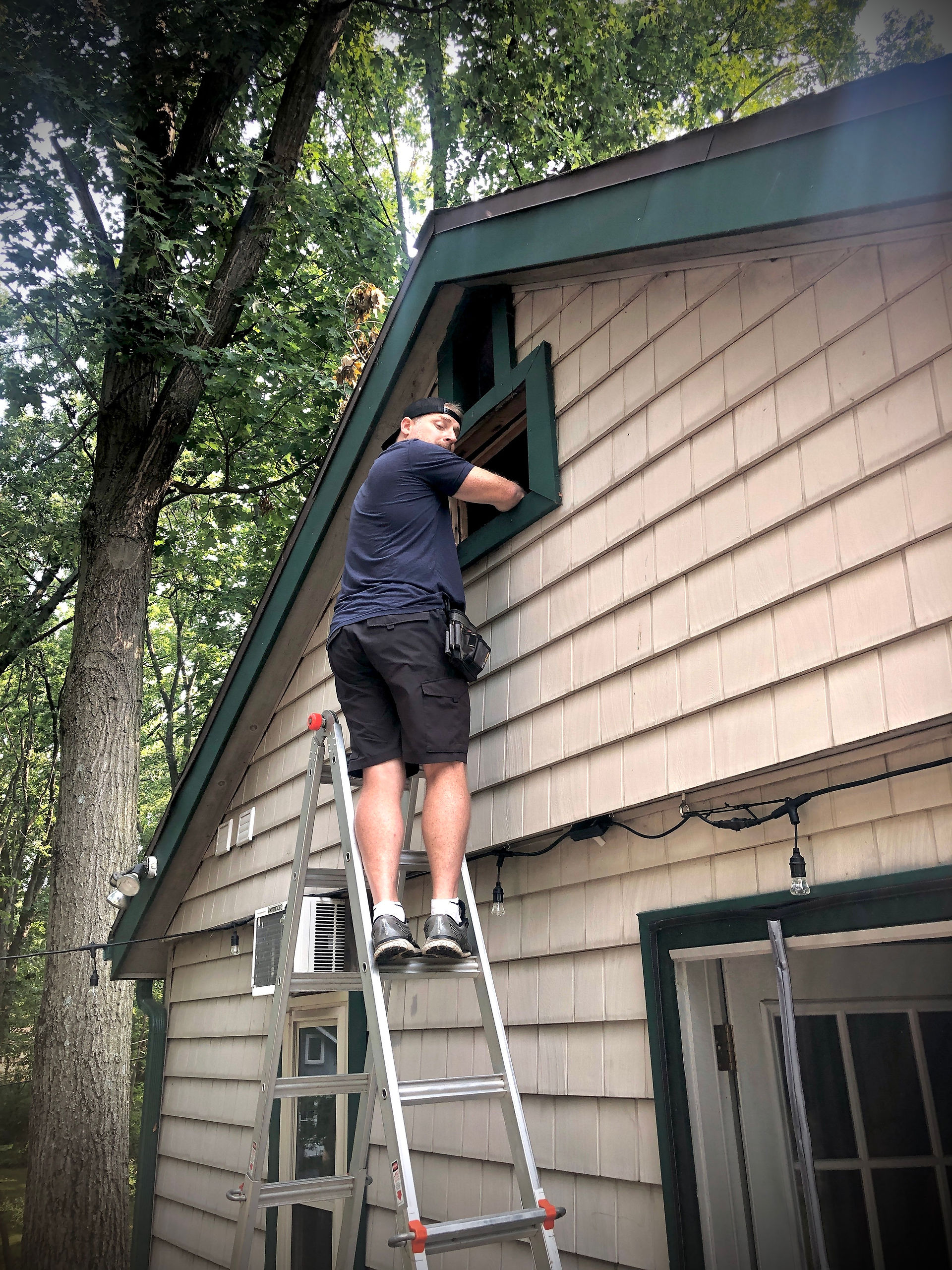 man standing on ladder removing window a/c unit from peak of house to prep for siding repair on lake house cottage