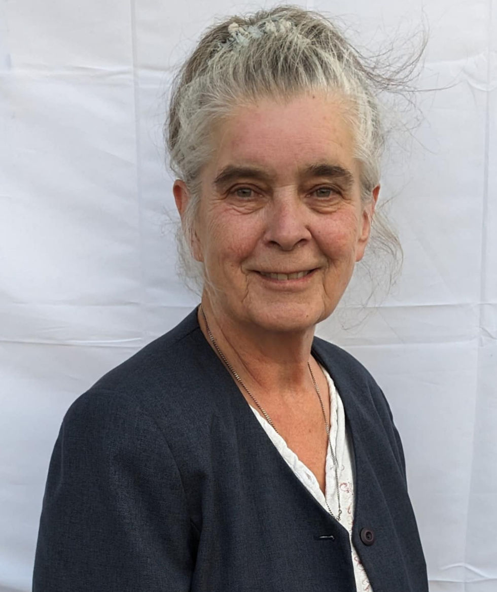 A woman with gray hair smiles gently, wearing a dark blazer over a white top, standing against a plain white background.
