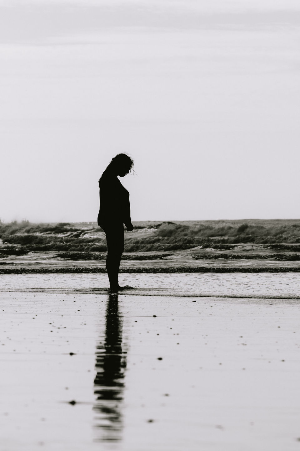 A lone person is captured in silhouette against the expansive backdrop of a beach, her reflection faintly mirrored on the wet sand. The image is a study in contrasts and simplicity, conveying a sense of peaceful isolation and the intimate connection we share with the natural world.