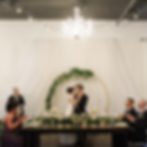 Couple at the head table under a gold arch with greenery and a chandelier at The Broz Event Venue